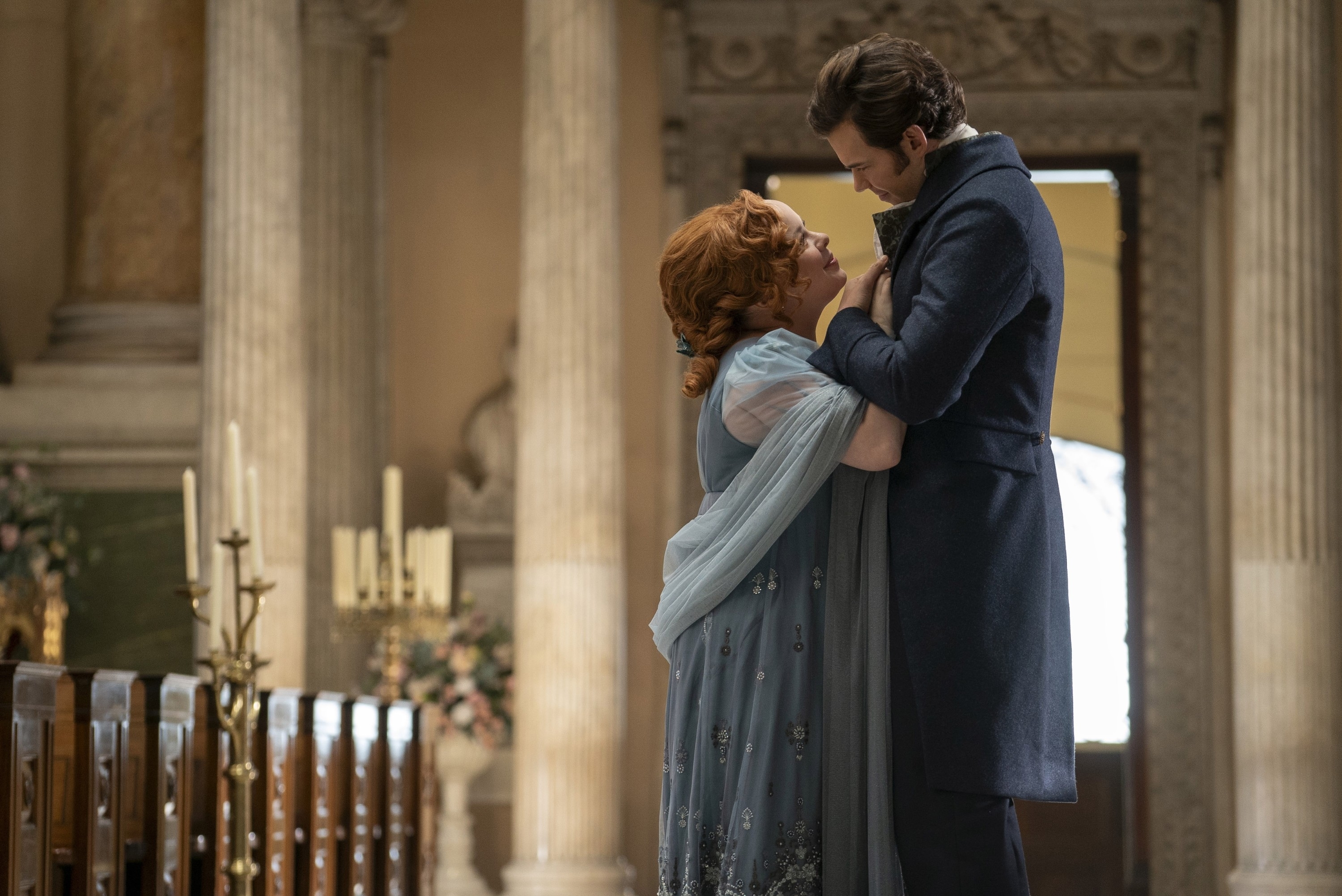 Nicola Coughlan in an elegant gown and Luke Newton in a formal coat embrace in a historical setting from the show "Bridgerton."