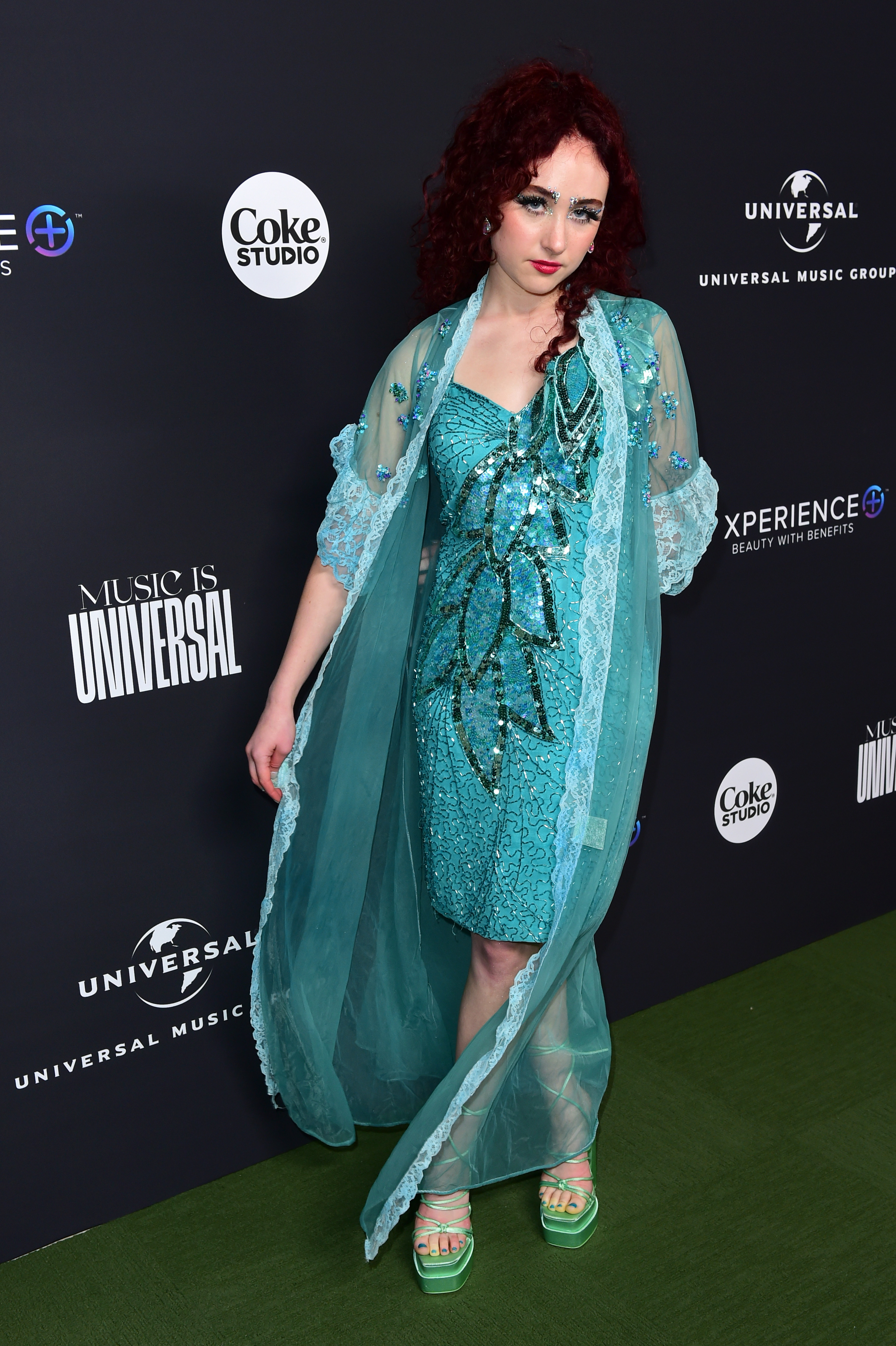 Young woman poses on red carpet in a glittery dress with leaf patterns and a sheer cape overlay. Text behind her includes &quot;Music is Universal&quot; and logos for Coke Studio and Universal Music Group