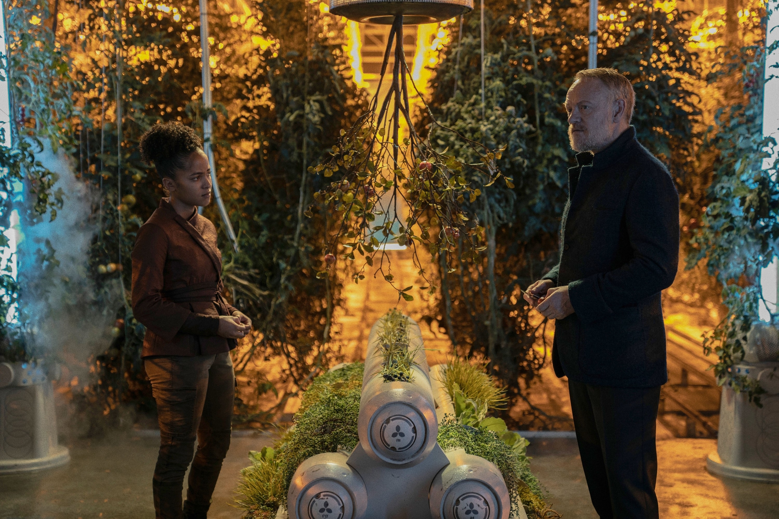 Jared Harris and Lou Llobell in a futuristic greenhouse scene from the TV series "Foundation," standing among tall plants and discussing