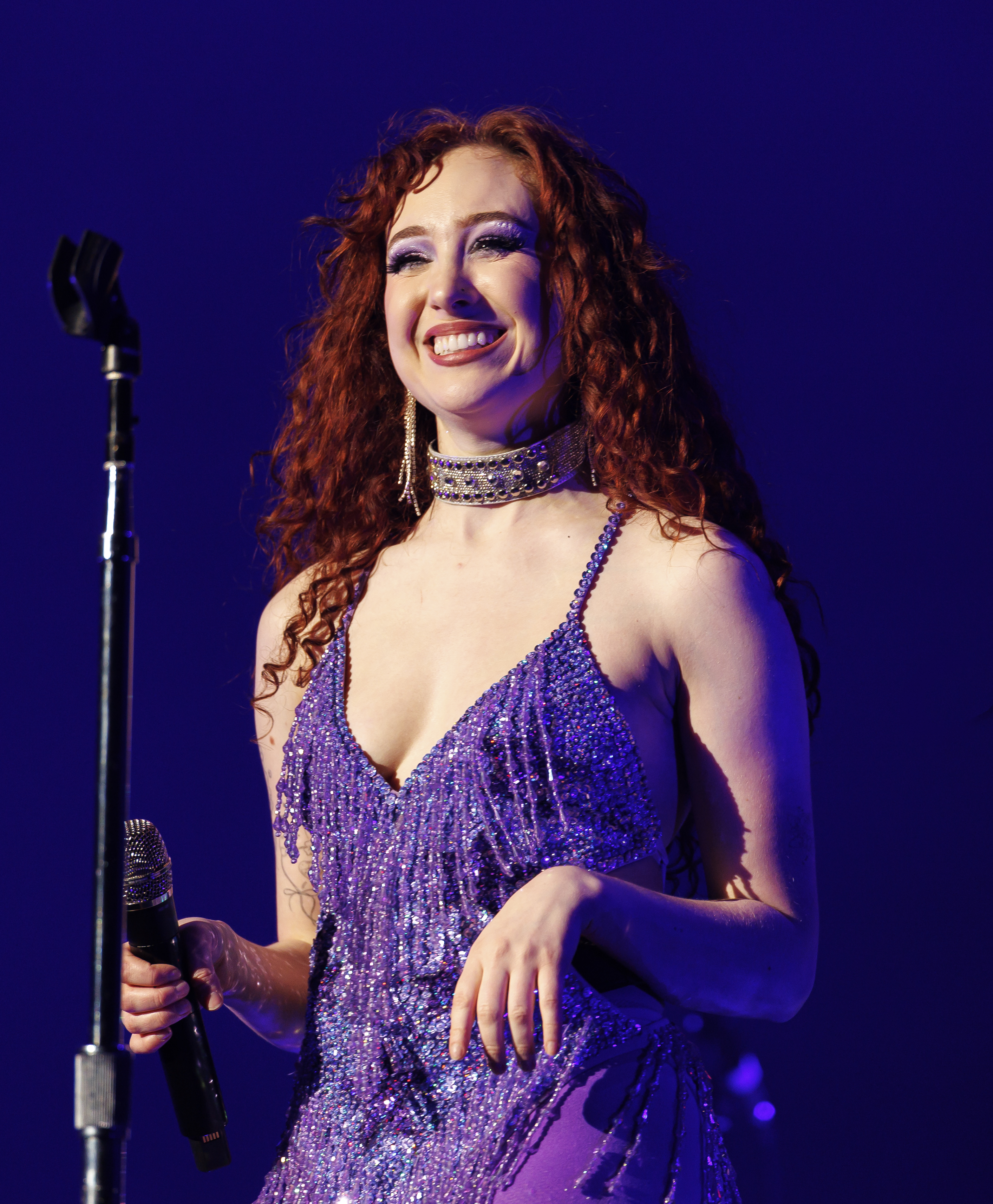 A singer with long red curly hair is smiling and holding a microphone on stage while wearing a sparkly dress with a choker