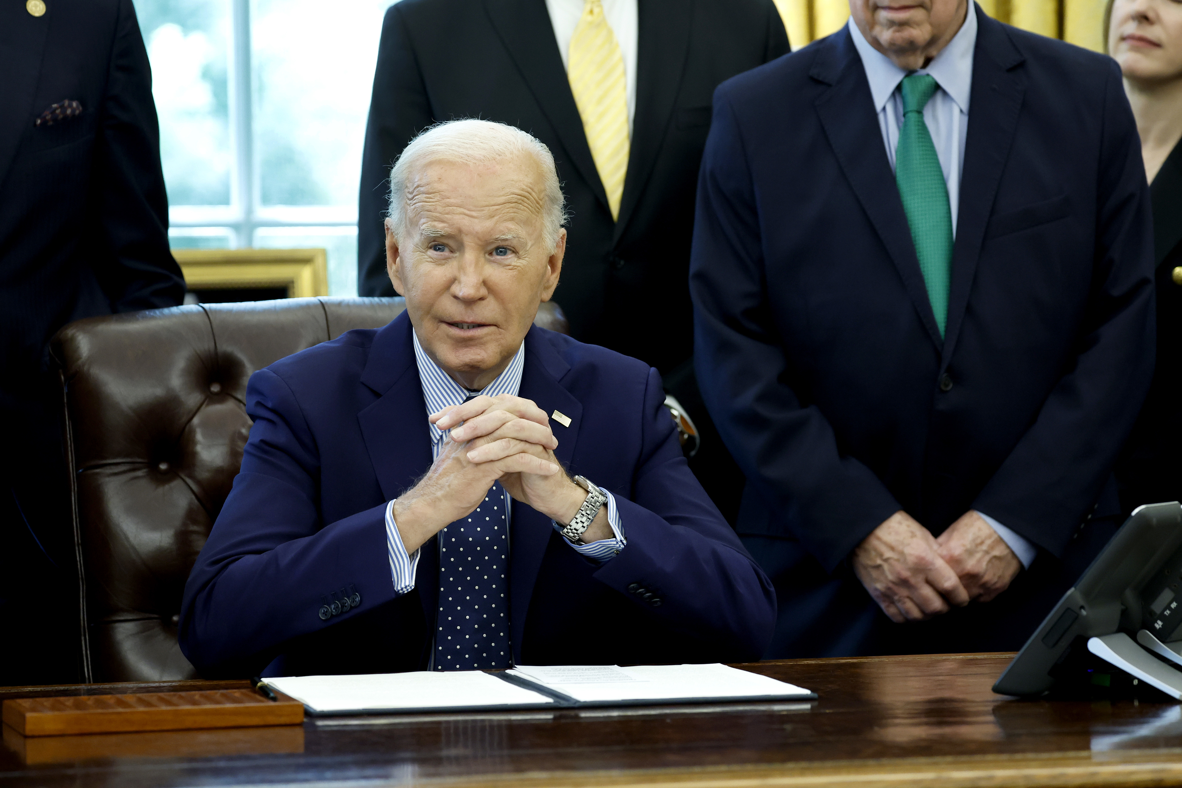 President Joe Biden sits at a desk in the Oval Office, surrounded by several standing men in suits