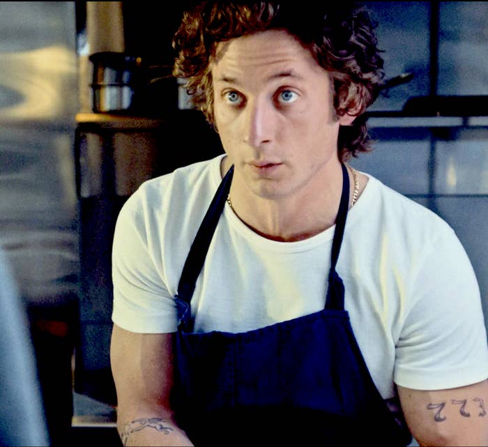 A man with curly hair, wearing a white shirt and a black apron, stands in a kitchen looking surprised. Tattoos are visible on his arms