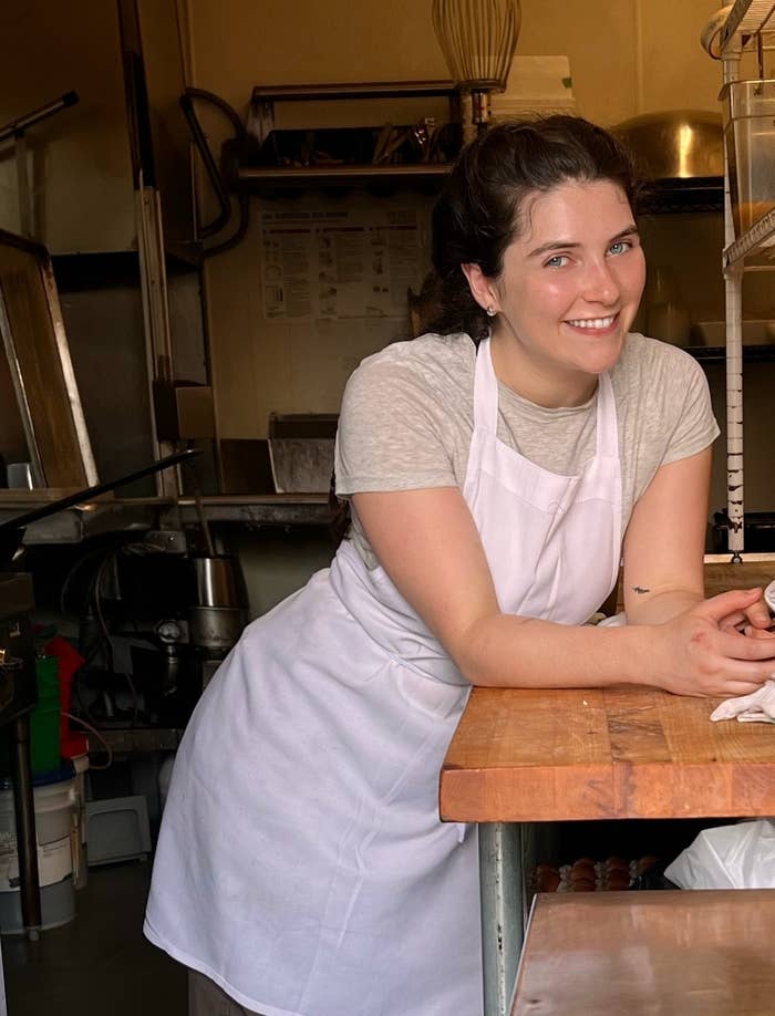 A person, likely a baker, stands leaning on a wooden counter in a bakery kitchen. Bread and baking ingredients are visible on the shelves around them