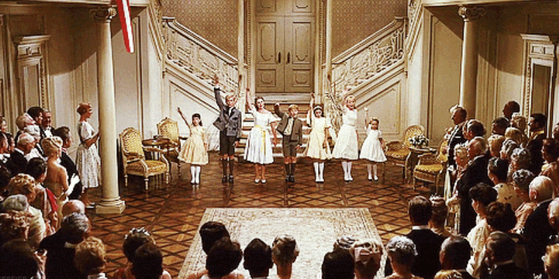 Six children are performing on stage in front of an audience in a large room with ornate decor. Four girls and two boys are holding hands, raising their arms