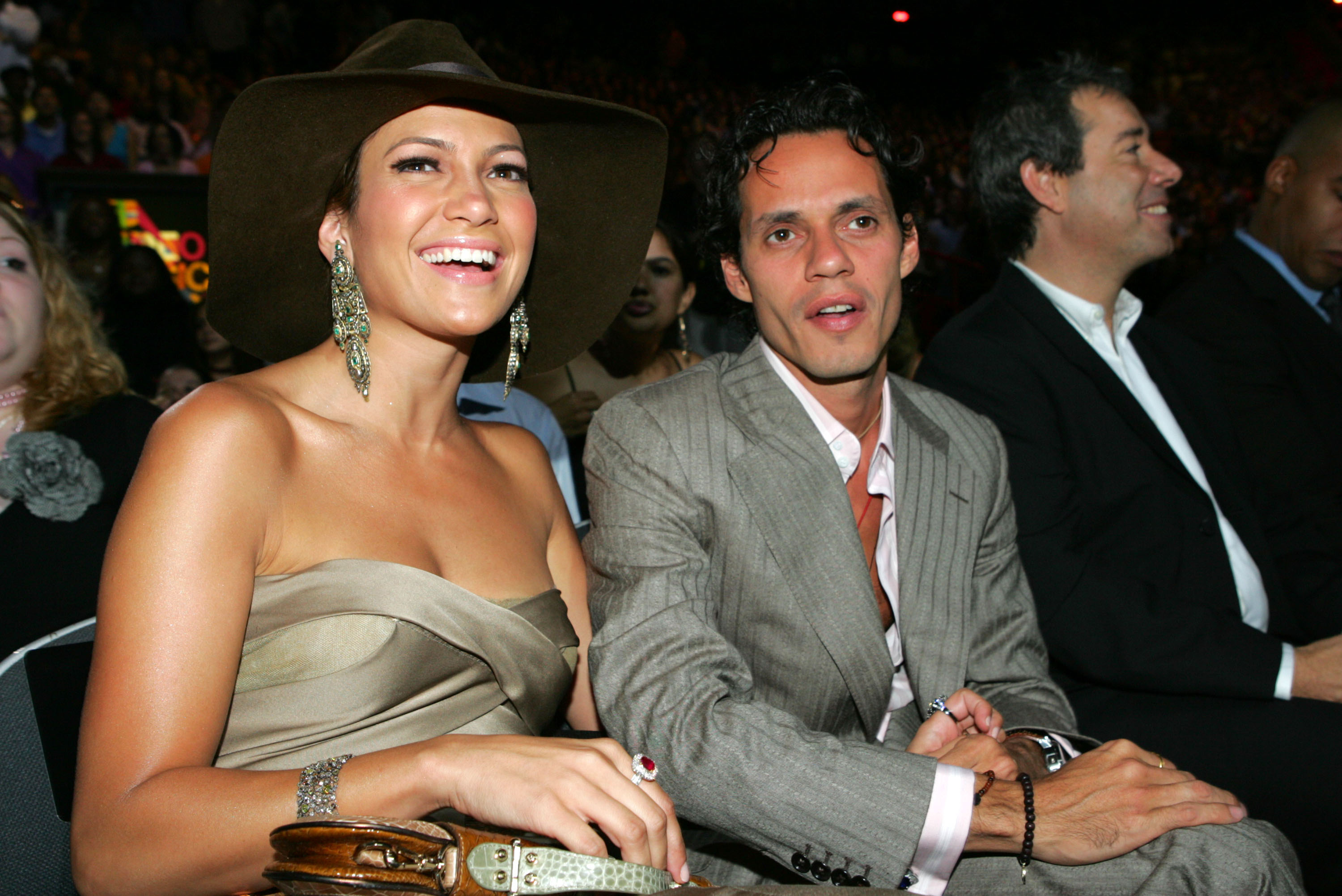 Jennifer Lopez wearing a strapless dress and wide-brim hat, sits next to Marc Anthony in a suit at an event, both smiling