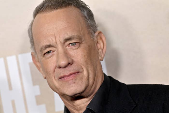 Tom Hanks in a close-up shot, wearing a dark suit jacket, against a blurred background with partial text