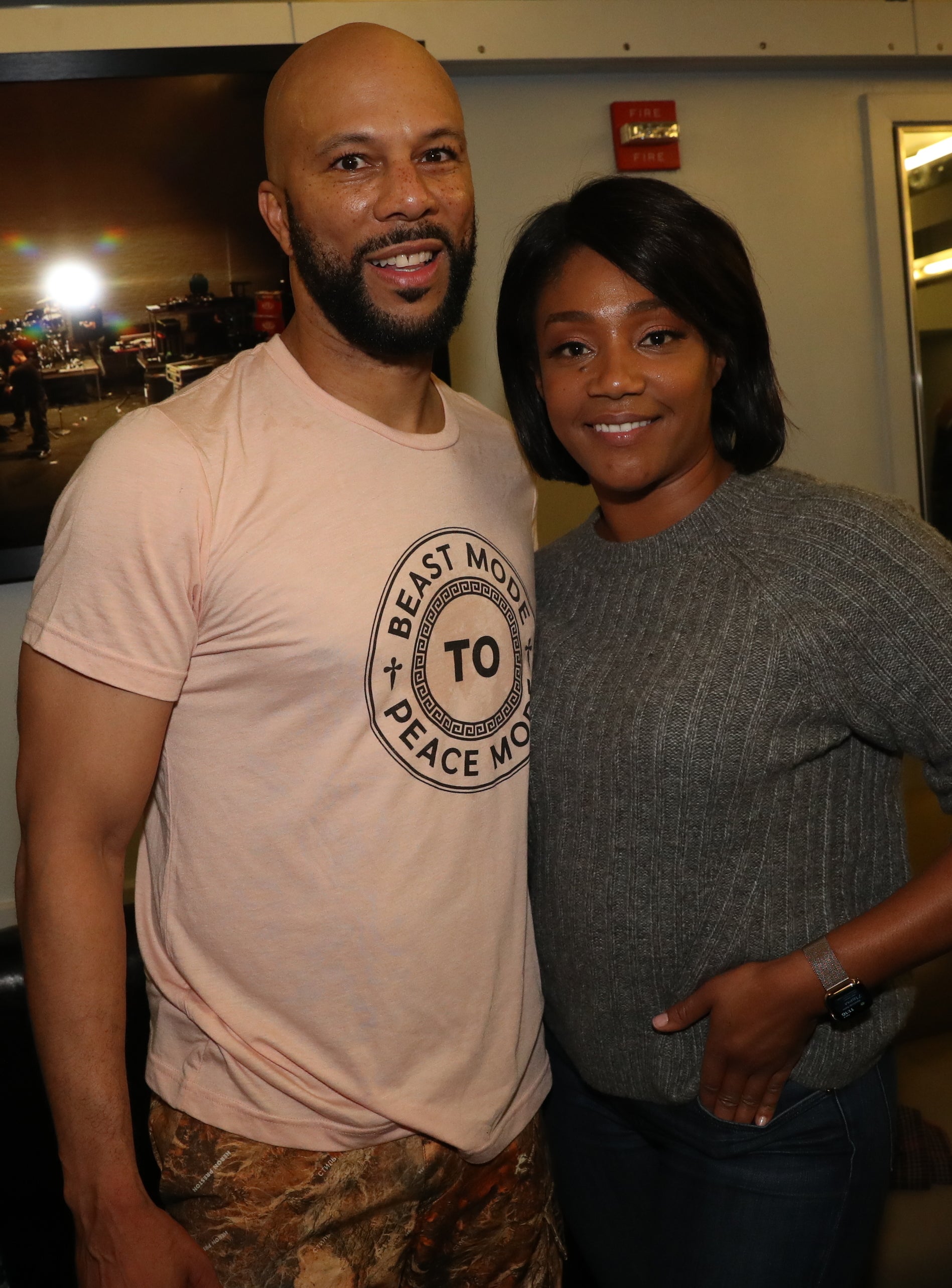 Common and Tiffany Haddish smiling and posing together indoors, with Common wearing a casual t-shirt and camouflage pants, and Tiffany in a sweater and jeans