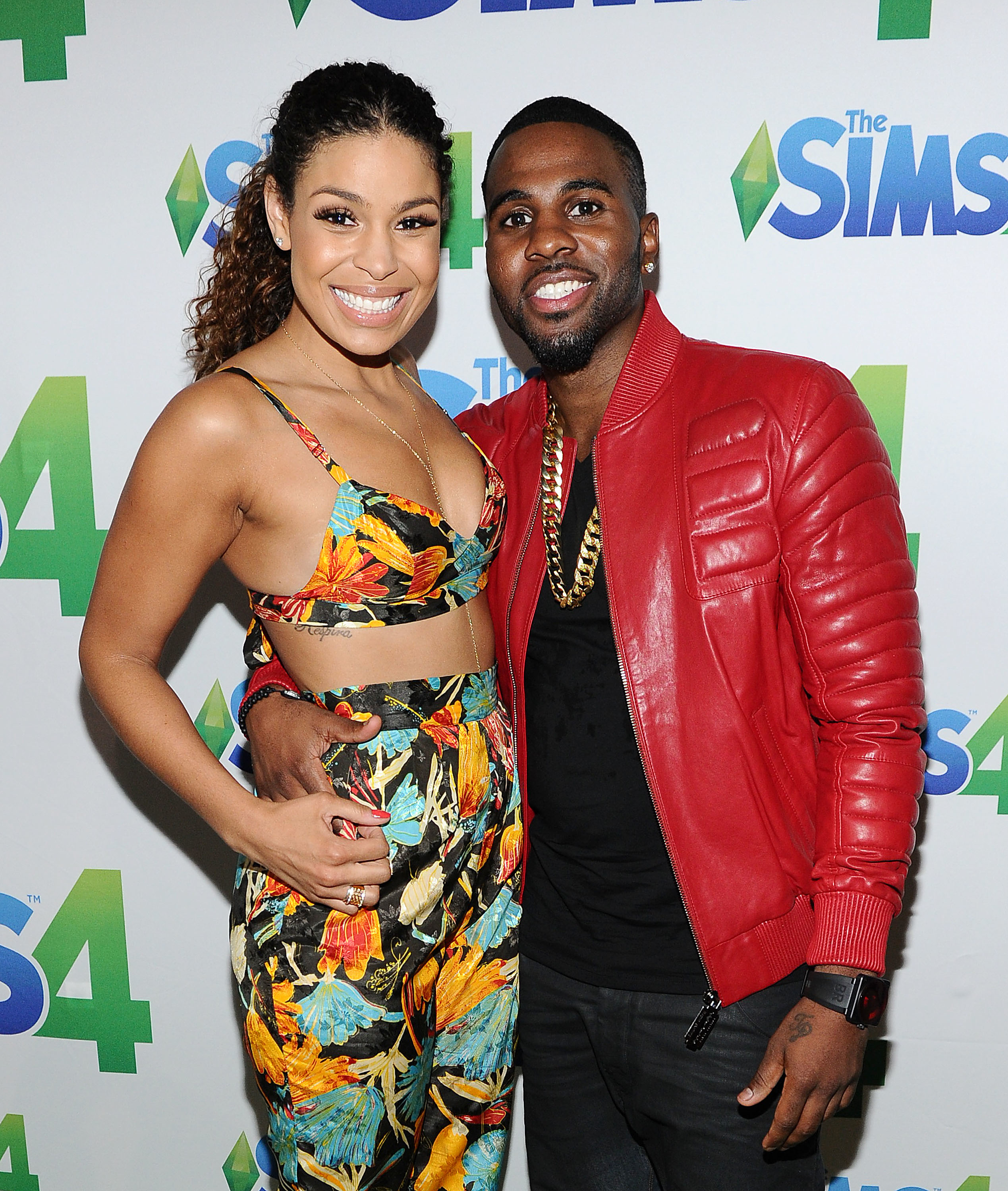 Jordin Sparks in a floral crop top and matching pants, stands with Jason Derulo in a red jacket at "The Sims 4" event backdrop, both smiling