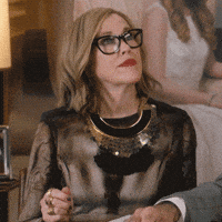 Catherine O'Hara, wearing glasses and a stylish black and sheer dress, is seen looking upwards