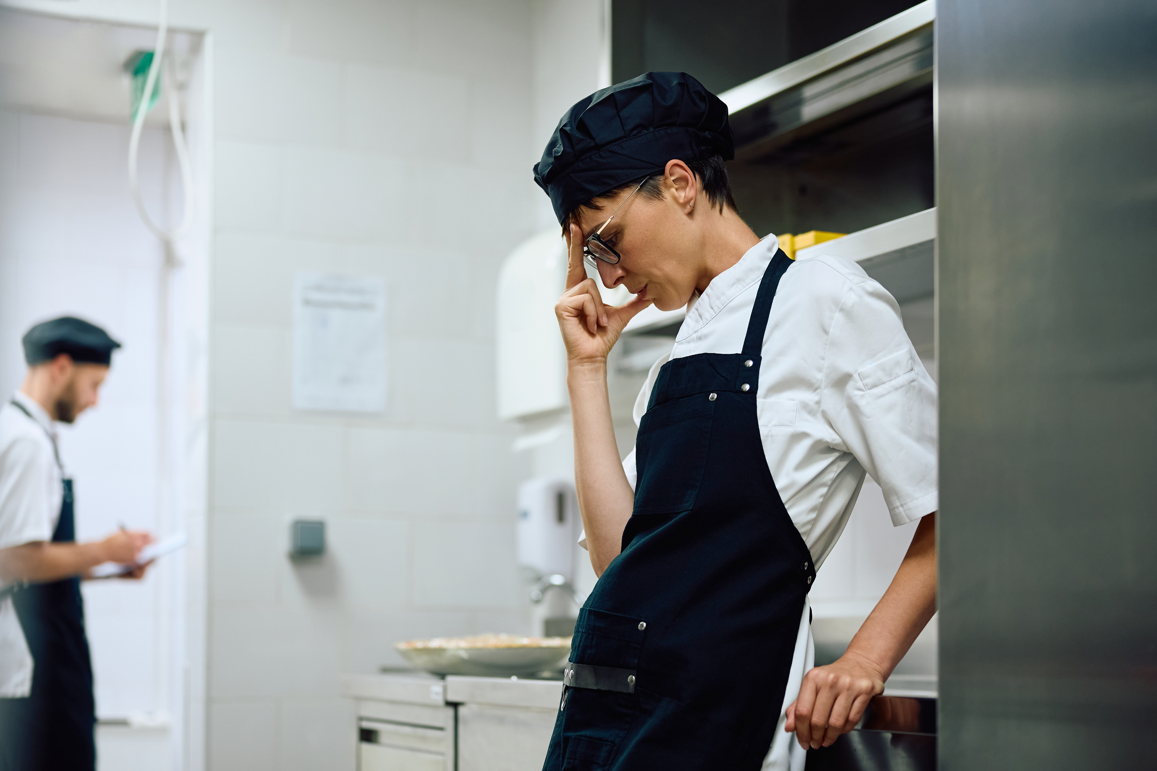 A chef in a professional kitchen wearing a white shirt, dark apron, and chef's hat leans against a counter, appearing pensive