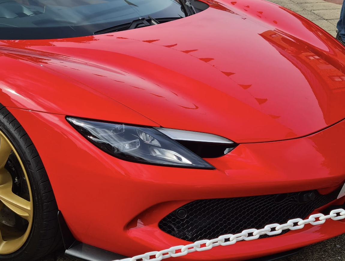 Close-up of the front of a red sports car with gold rims, showcasing the sleek design and detailing. A white chain barrier is in front of the car