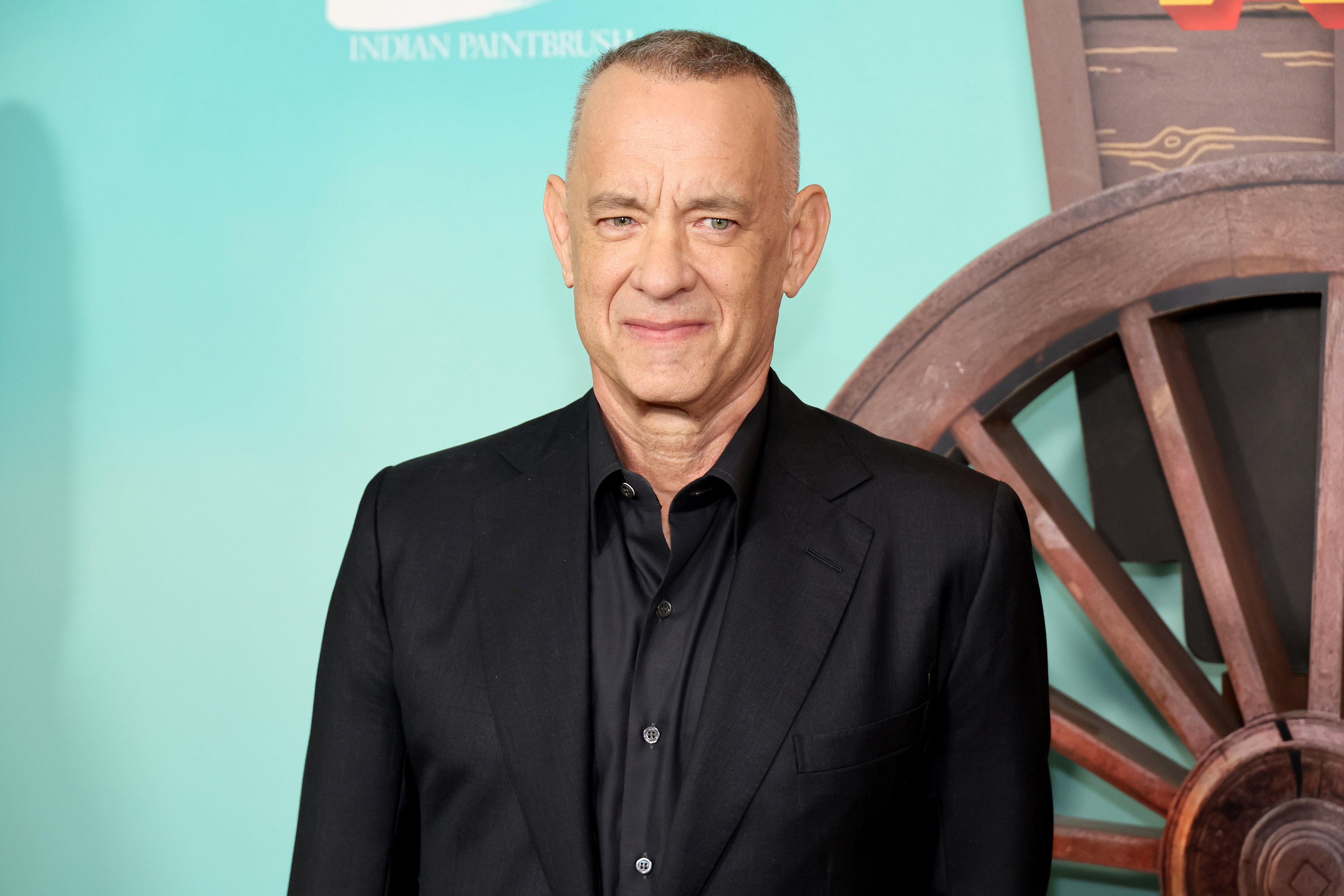 Tom Hanks smiles at an event in formal attire with a background including a wagon wheel