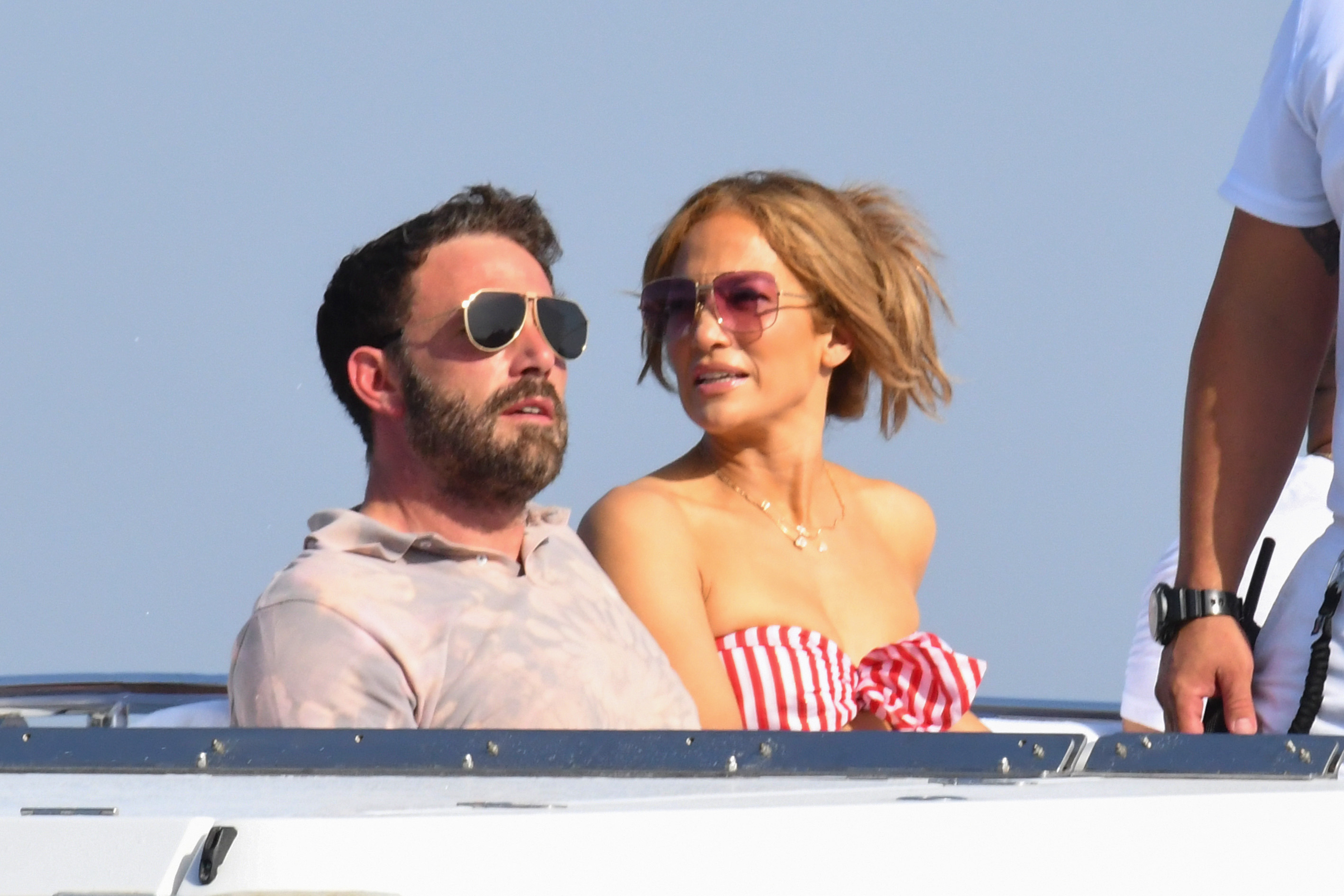 Ben Affleck and Jennifer Lopez are relaxing on a boat. Ben is wearing a casual shirt, and Jennifer is in a strapless outfit with sunglasses