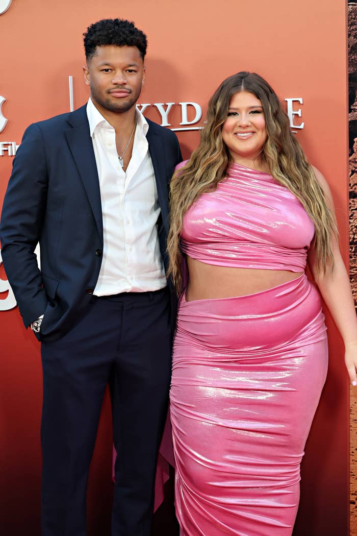 Chase Stokes in a navy suit and Madelyn Cline in a pink gown are posing together on the red carpet