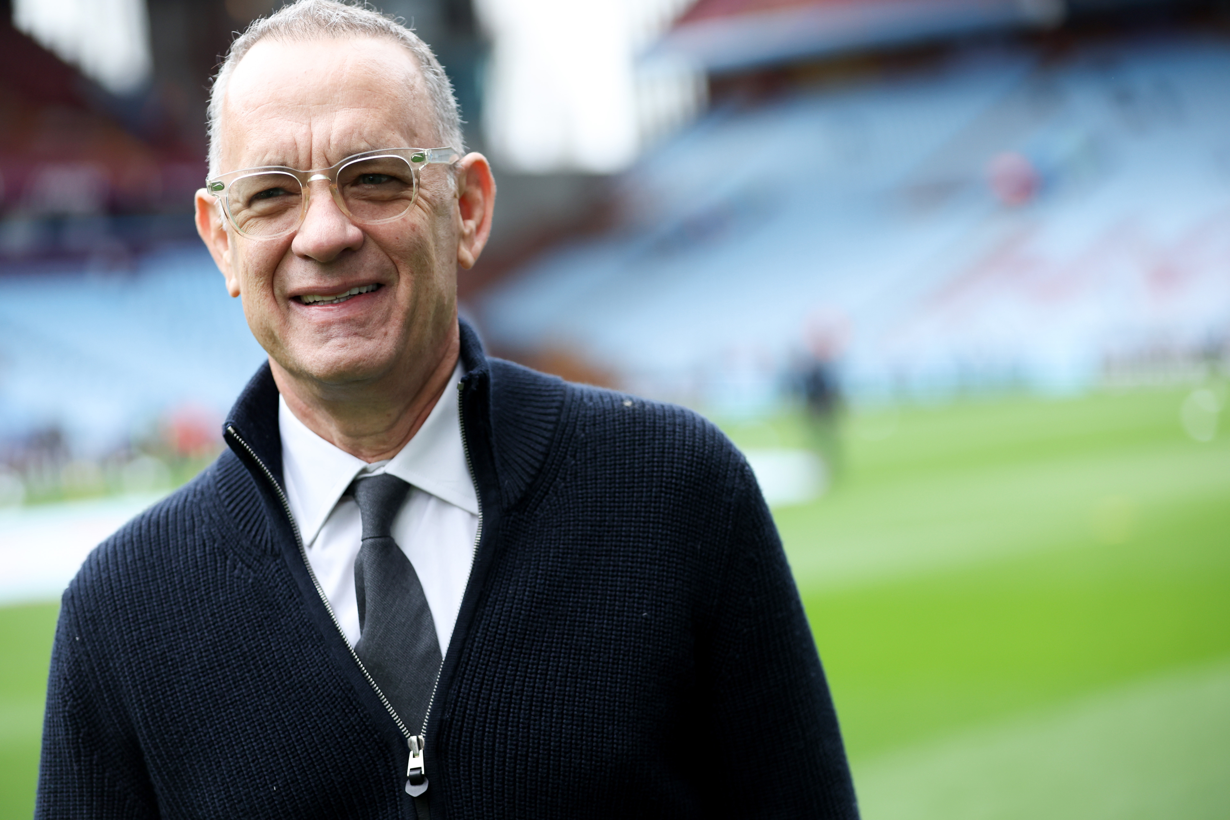 Tom Hanks is smiling while standing in a sports stadium. He is wearing glasses, a zip-up sweater, a shirt, and a tie