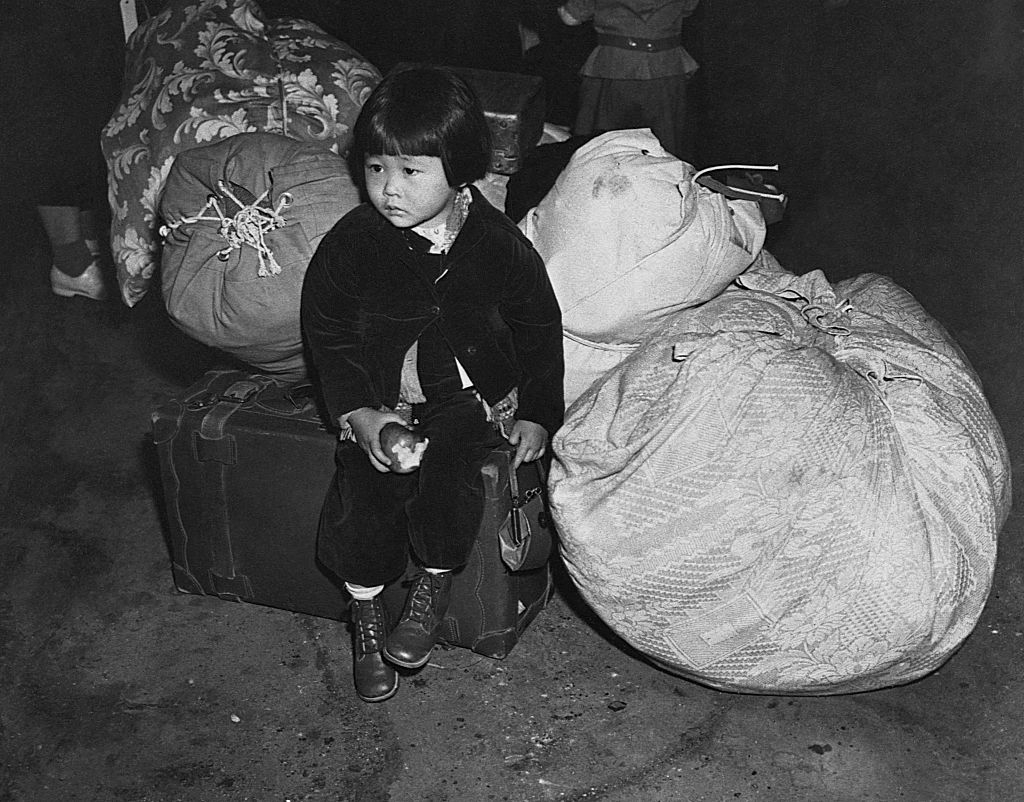 A young child sits on a suitcase, holding an item, with large tied bundles in the background. The image has a historical feel