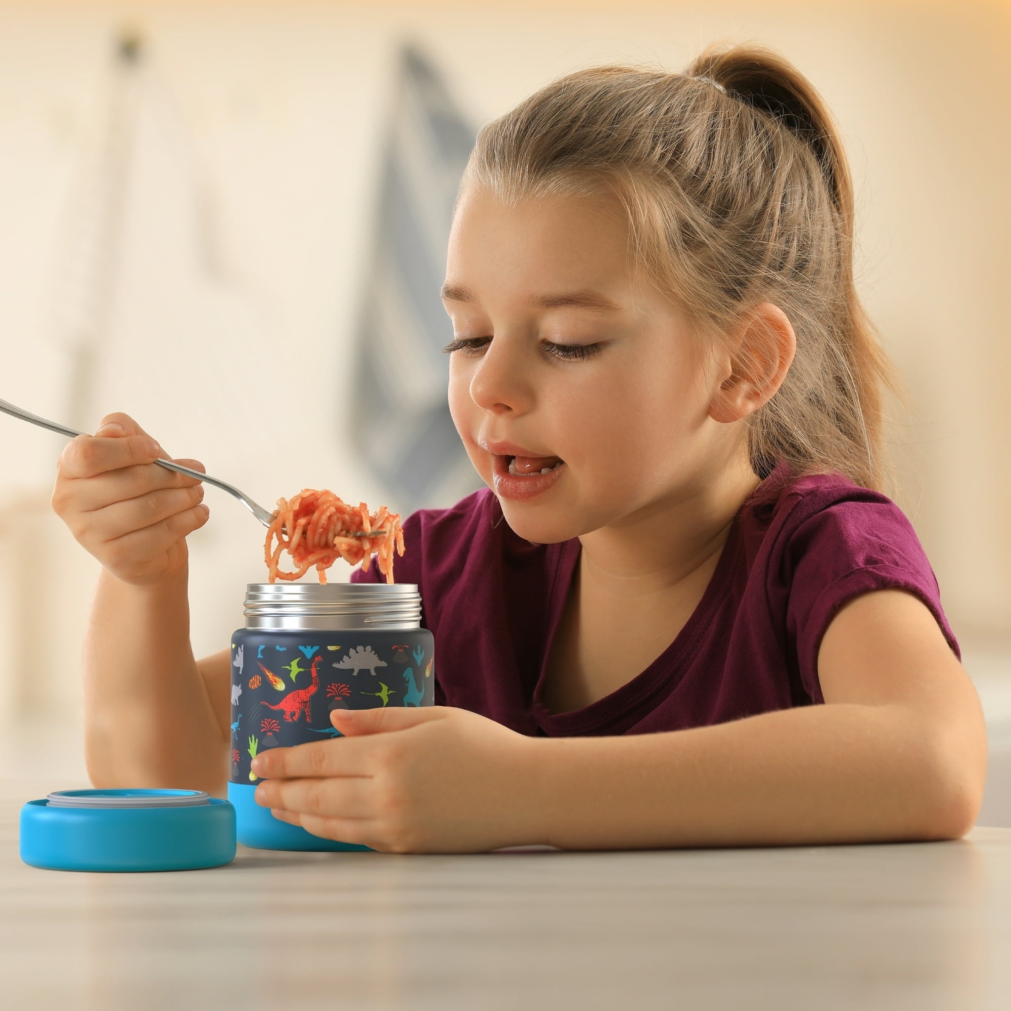 A child in a casual outfit is eating noodles from a thermal container with a colorful dinosaur design