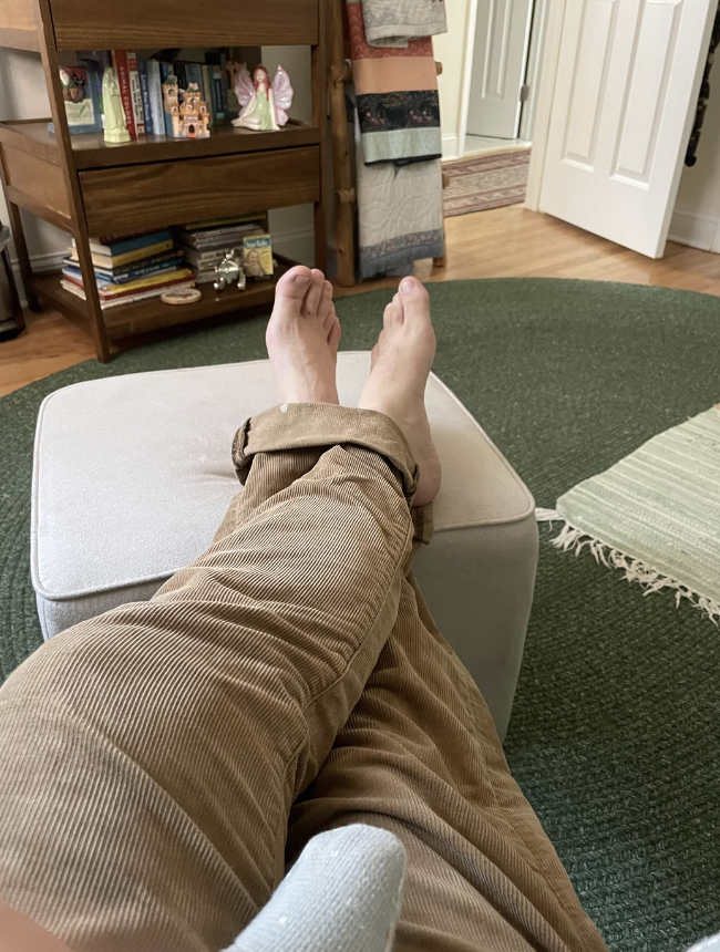 A person with rolled-up pants is relaxing with their feet up on an ottoman in a cozy, homey room with bookshelves and carpets