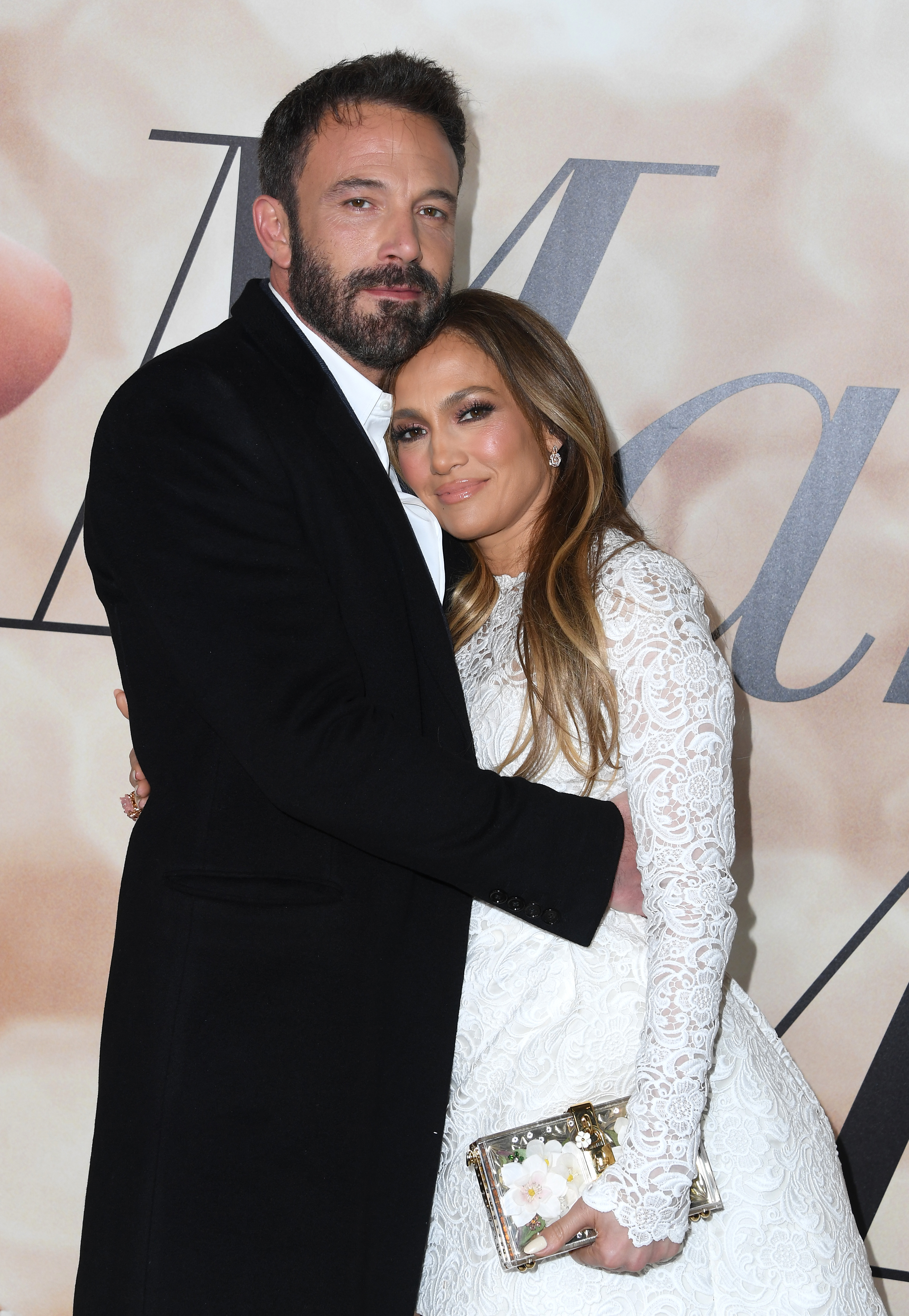 Ben Affleck and Jennifer Lopez pose together; Ben is in a dark suit and Jennifer wears a white lace dress, holding a floral clutch