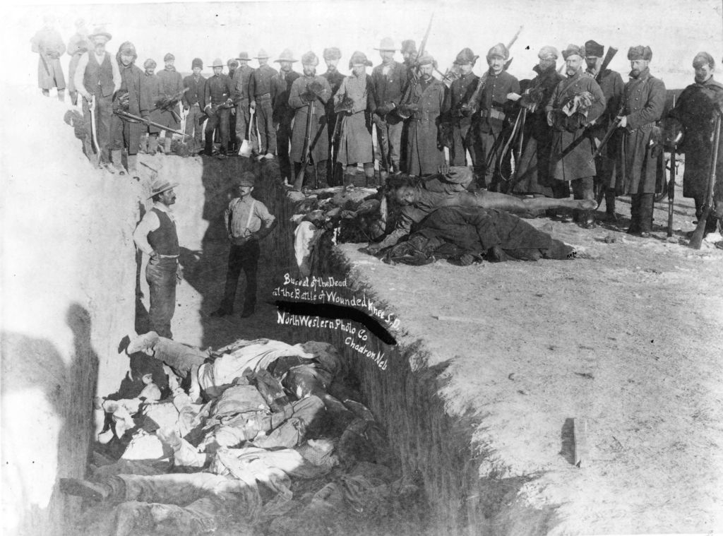 Historical photograph of the Wounded Knee Massacre, showing victims in a mass grave and armed soldiers standing at the edge
