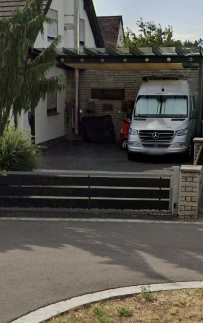 A modern driveway with a Mercedes-Benz van and a red scooter parked under a carport attached to a house, surrounded by trees and shrubbery