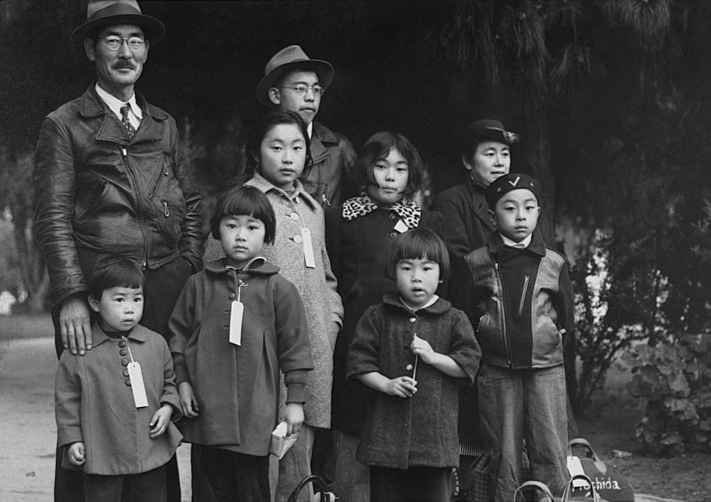 A historical family photo shows seven children and two adults, all wearing numbers tags, in front of a dark background