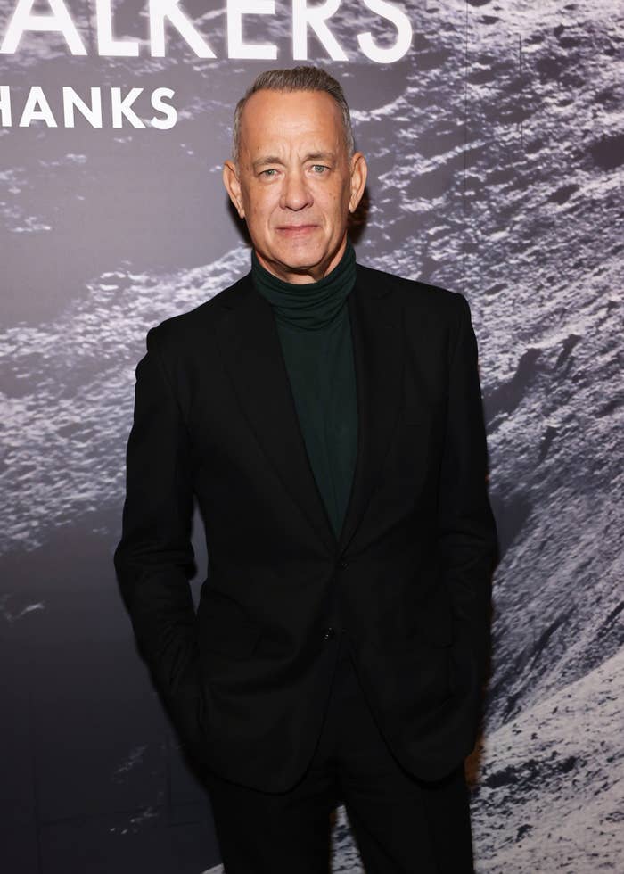 Tom Hanks stands in front of a textured backdrop, wearing a dark suit with a dark turtleneck