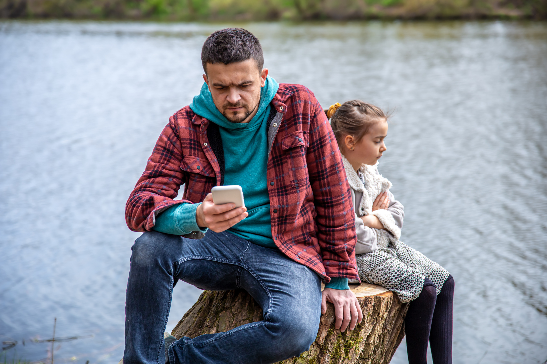 An adult man and a young girl sit by a lake. The man, in casual attire, looks at his phone while the girl sits with her arms crossed, looking away