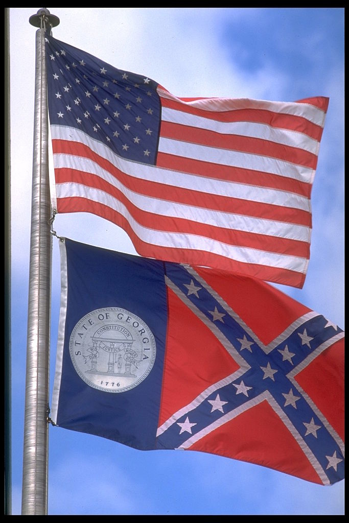 An American flag and an older version of the Georgia state flag with the Confederate battle emblem are flying on a flagpole against a clear sky