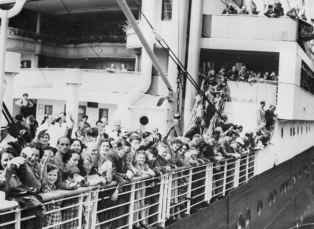 Large crowd of people, many of whom are smiling and waving, stand on the deck of a ship as it docks, suggesting a significant and historic event
