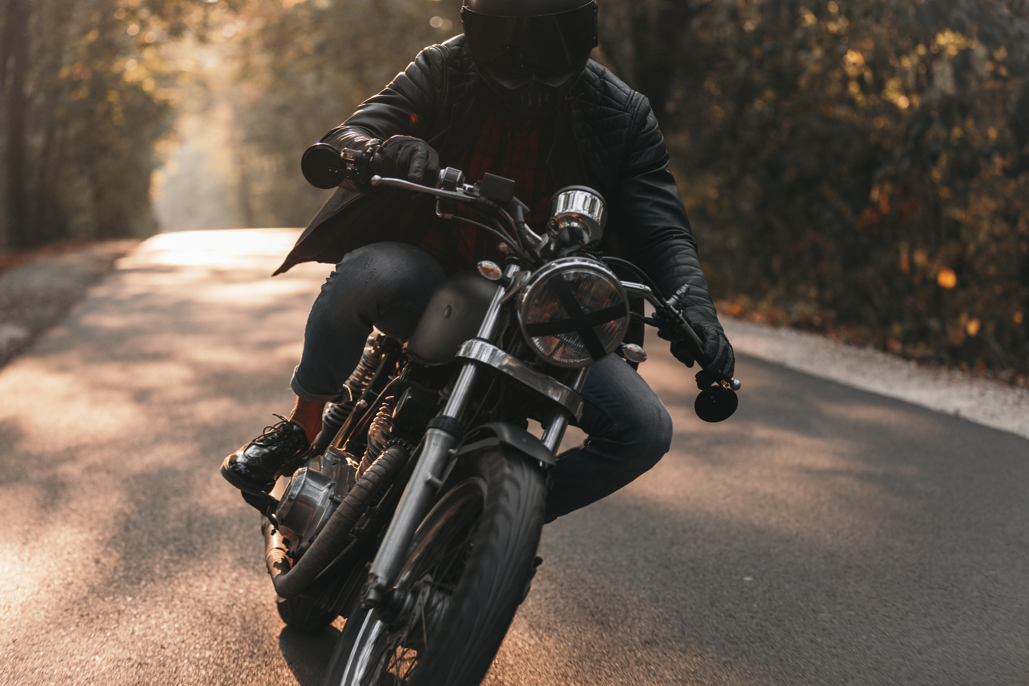 Person riding a motorcycle on a sunlit road, wearing a helmet, leather jacket, jeans, and boots