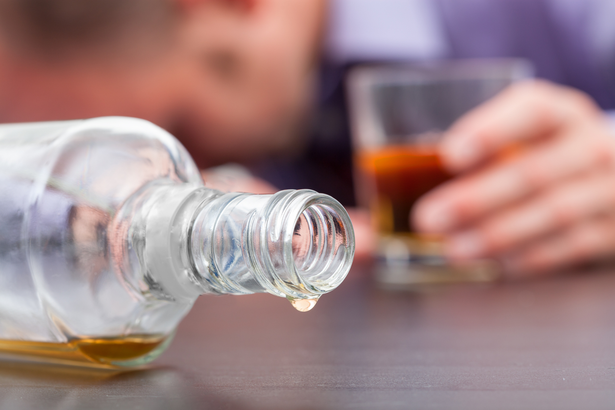 Close-up of a person holding a drink with their head down on a table beside a nearly empty bottle of alcohol