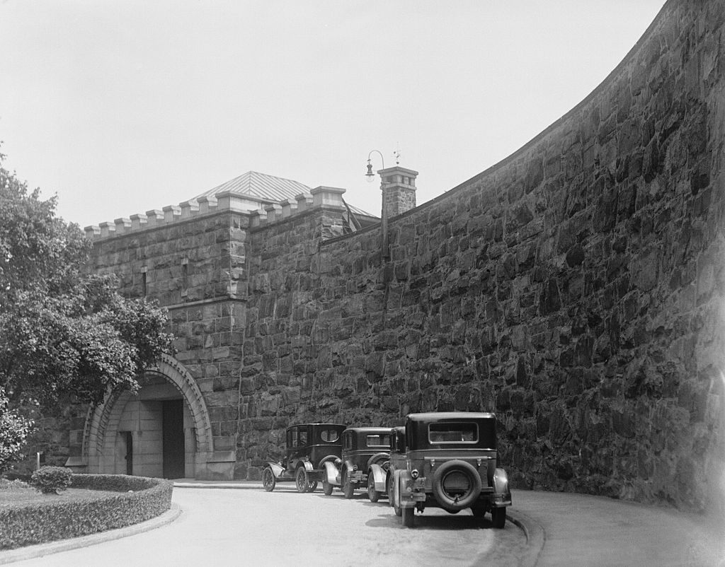 Historic photo of three vintage cars driving past a tall stone wall near the entrance of a castle-like building