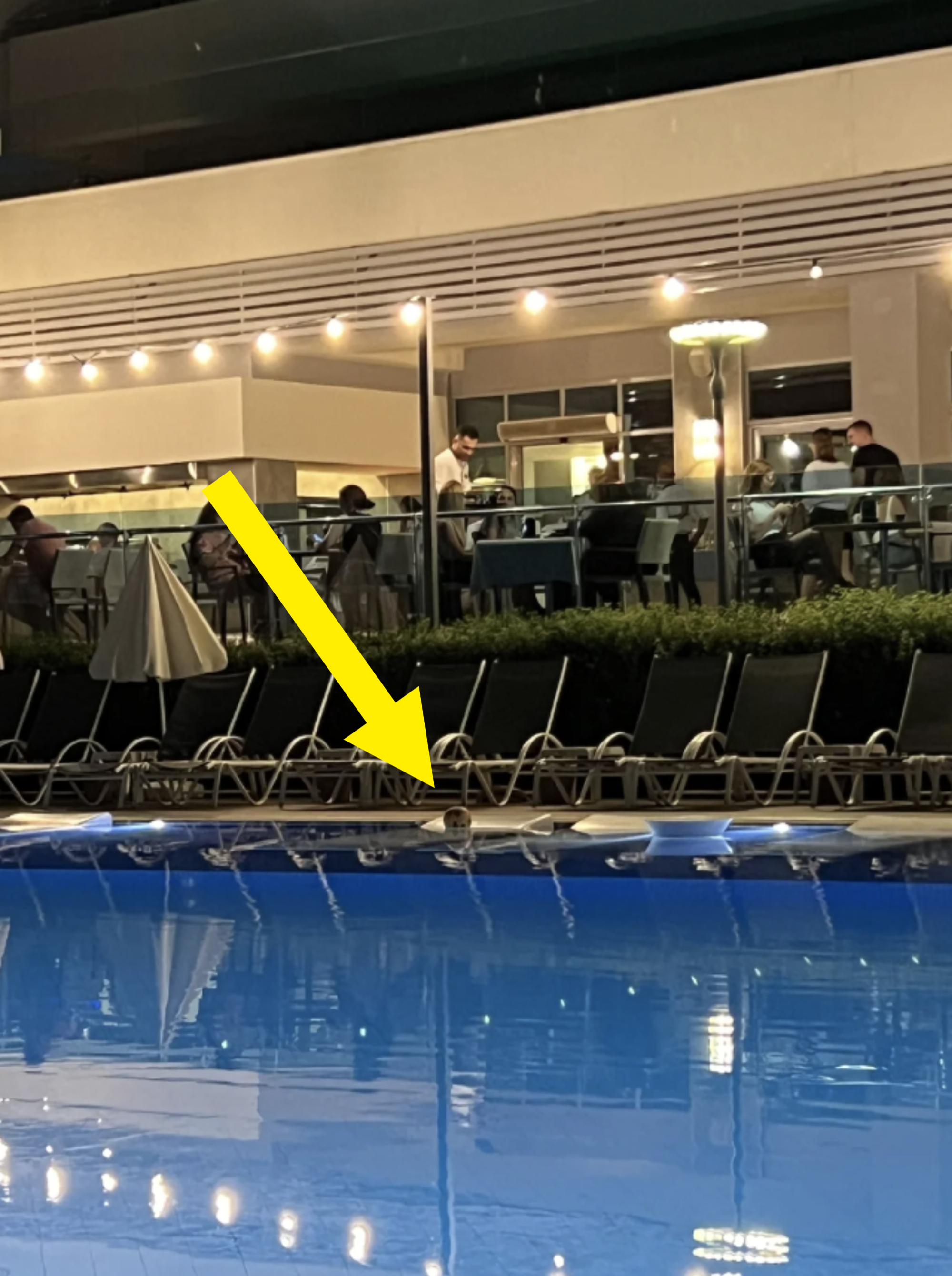 People dining at a poolside restaurant in the evening, with the pool in the foreground and a modern building's terrace in the background