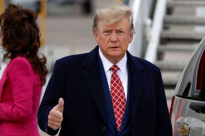 Donald Trump gives a thumbs-up gesture while outside, with a person in the background. He is wearing a suit with a patterned red tie