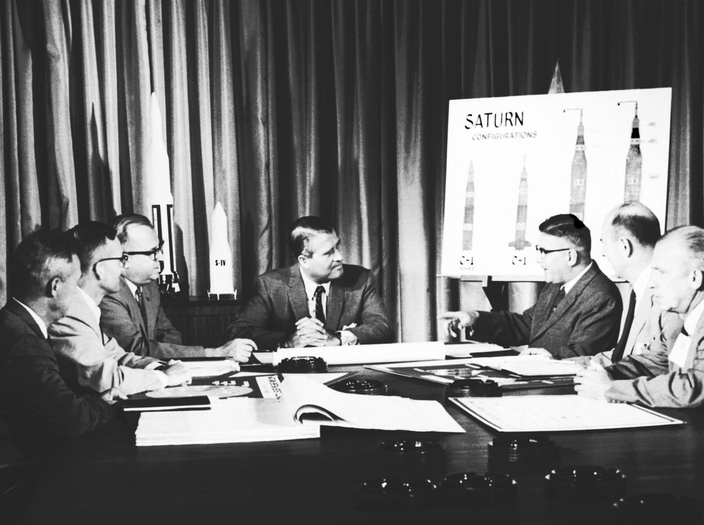 Group of men, including Wernher von Braun, sit around a table discussing a chart of Saturn rocket comparisons displayed on an easel