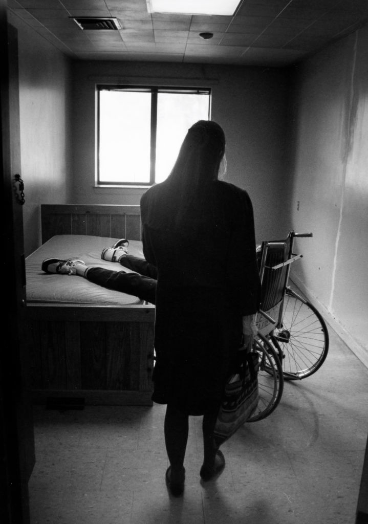 A woman stands in a small room, looking at a person lying on a bed with their legs extended and a wheelchair beside the bed