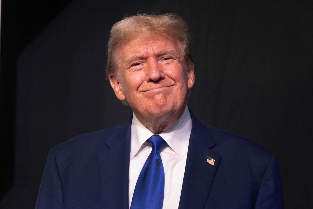Donald Trump in a suit with a tie, slightly smiling, with an American flag pin on his lapel