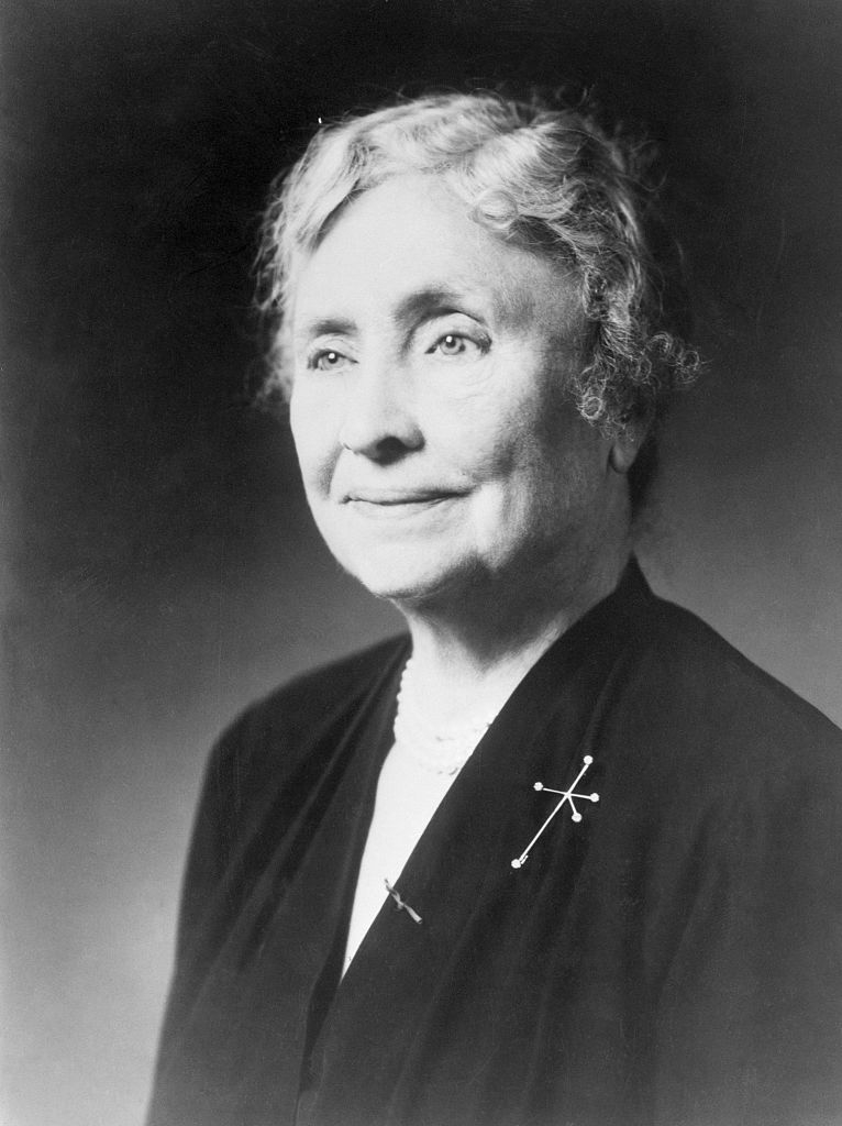 Helen Keller, in a formal portrait wearing a dark dress adorned with simple jewelry, smiles gently