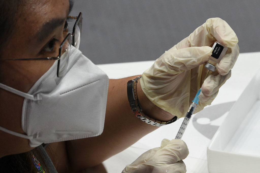 A person wearing glasses and a mask is preparing a syringe with a vaccine from a vial
