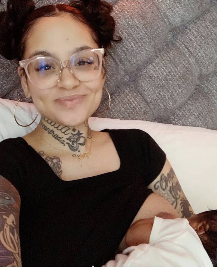 A smiling woman with tattoos and glasses, wearing a black top, is breastfeeding a baby. The woman has her hair styled in two buns