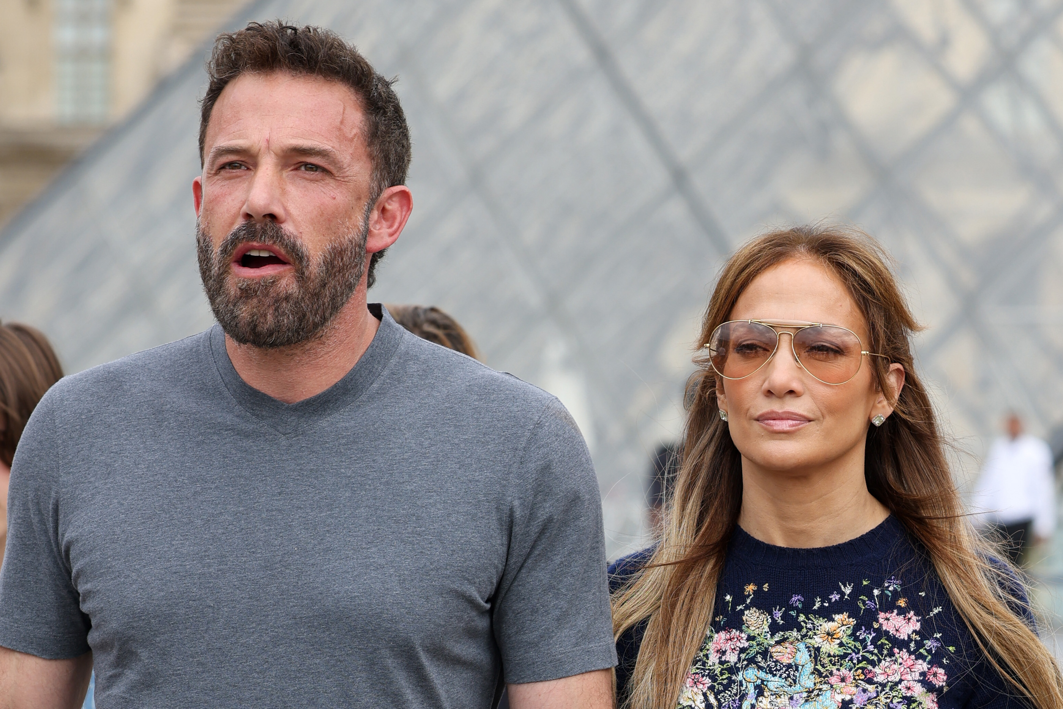 Ben Affleck and Jennifer Lopez walk together outdoors. Affleck wears a t-shirt, and Lopez wears a top adorned with a floral design and sunglasses