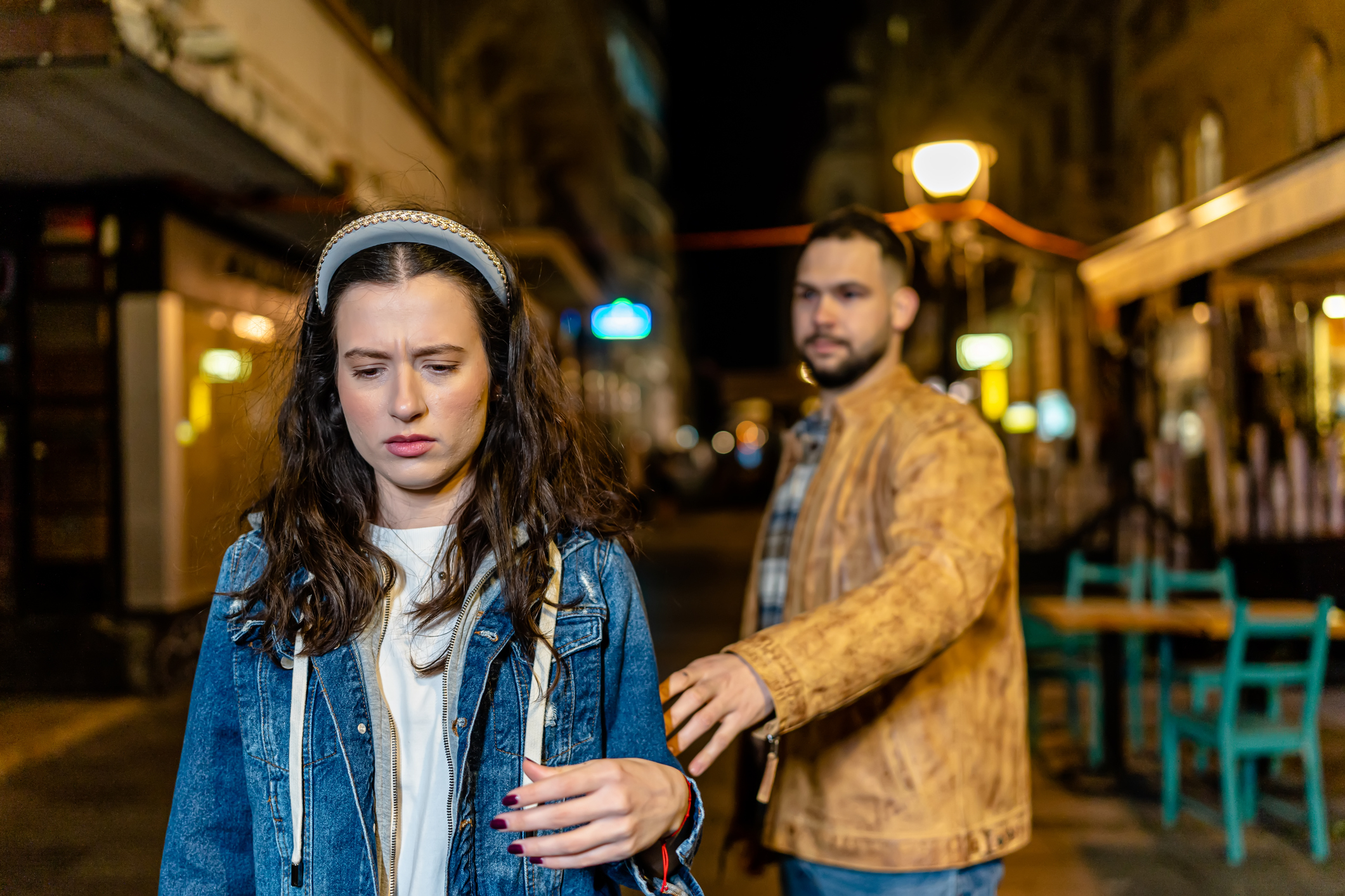 Woman in denim jacket walks away looking upset, while a man in a brown jacket reaches out to her on a dimly lit street