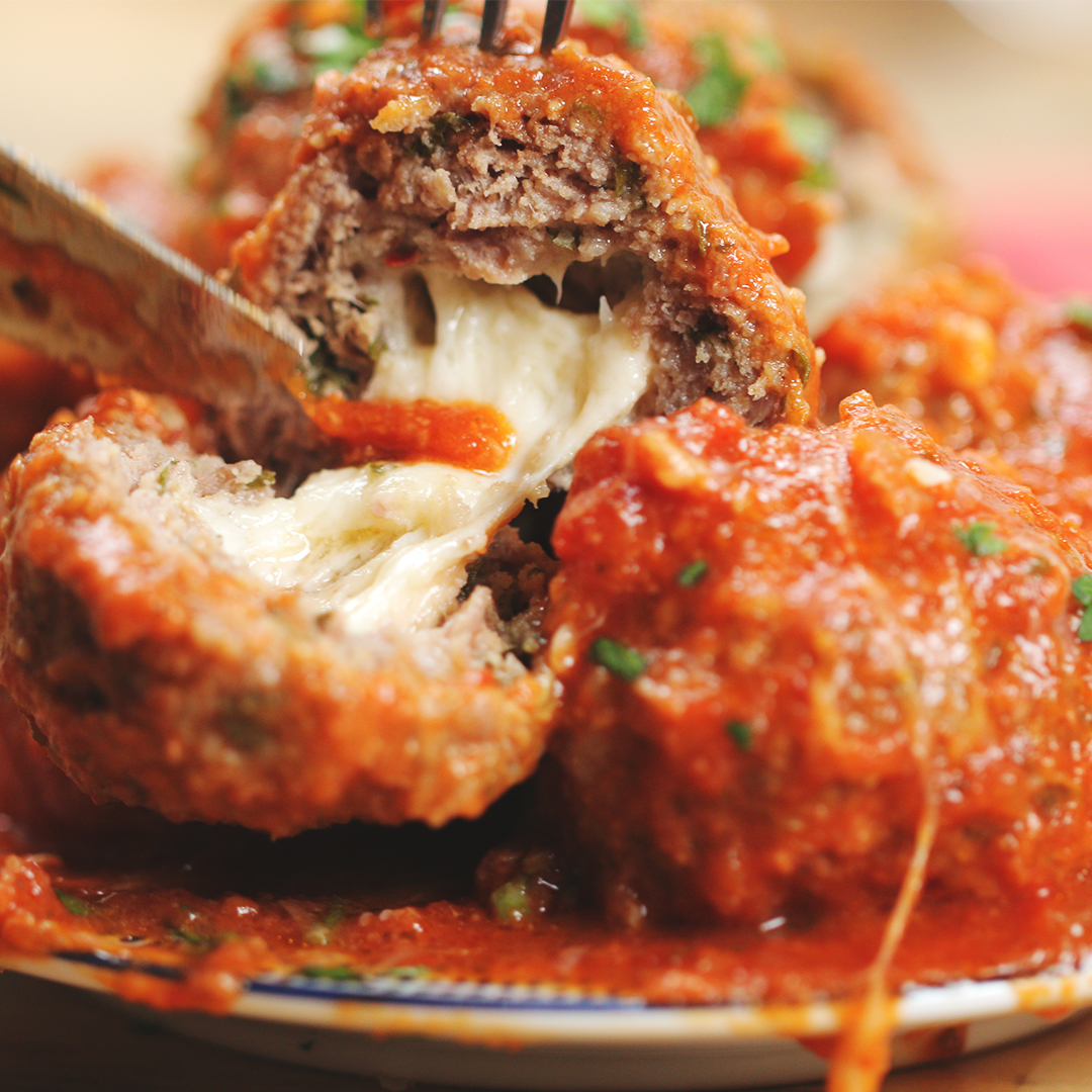 A plate of meatballs stuffed with melted cheese, topped with tomato sauce, being sliced open with a fork and knife