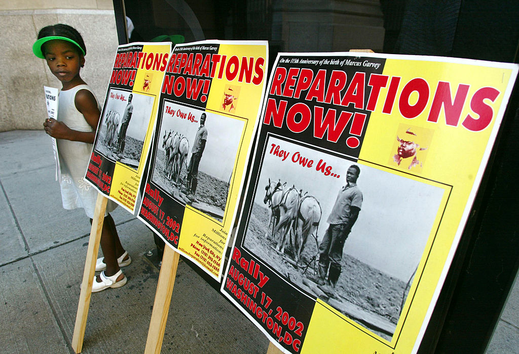 A young girl stands next to signs that read &quot;Reparations Now! They Owe Us&hellip; Rally August 17, 2002, Washington D.C.&quot;