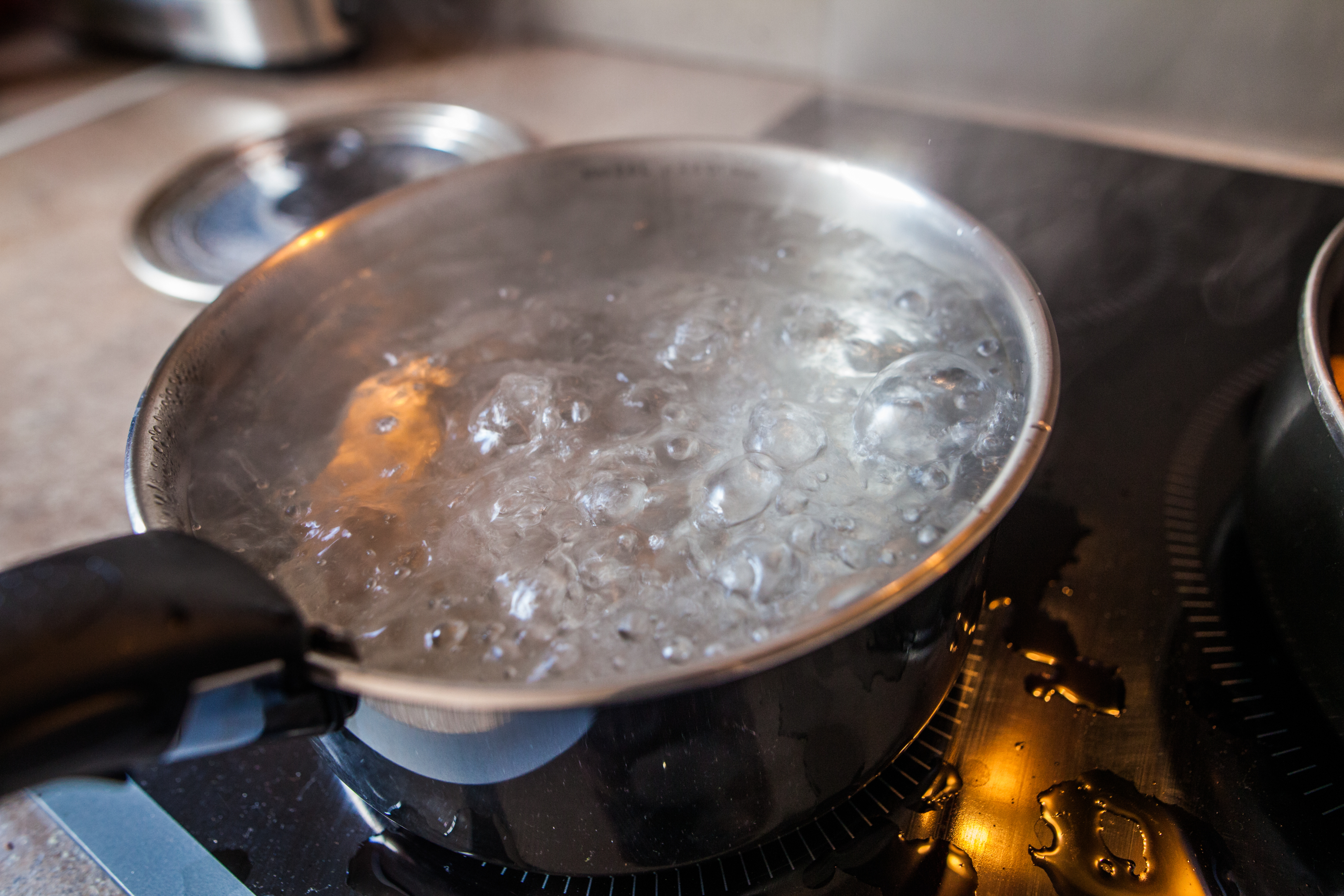 A pot of water is boiling on a stovetop, with bubbles rising to the surface. Another pot with a lid is visible in the background