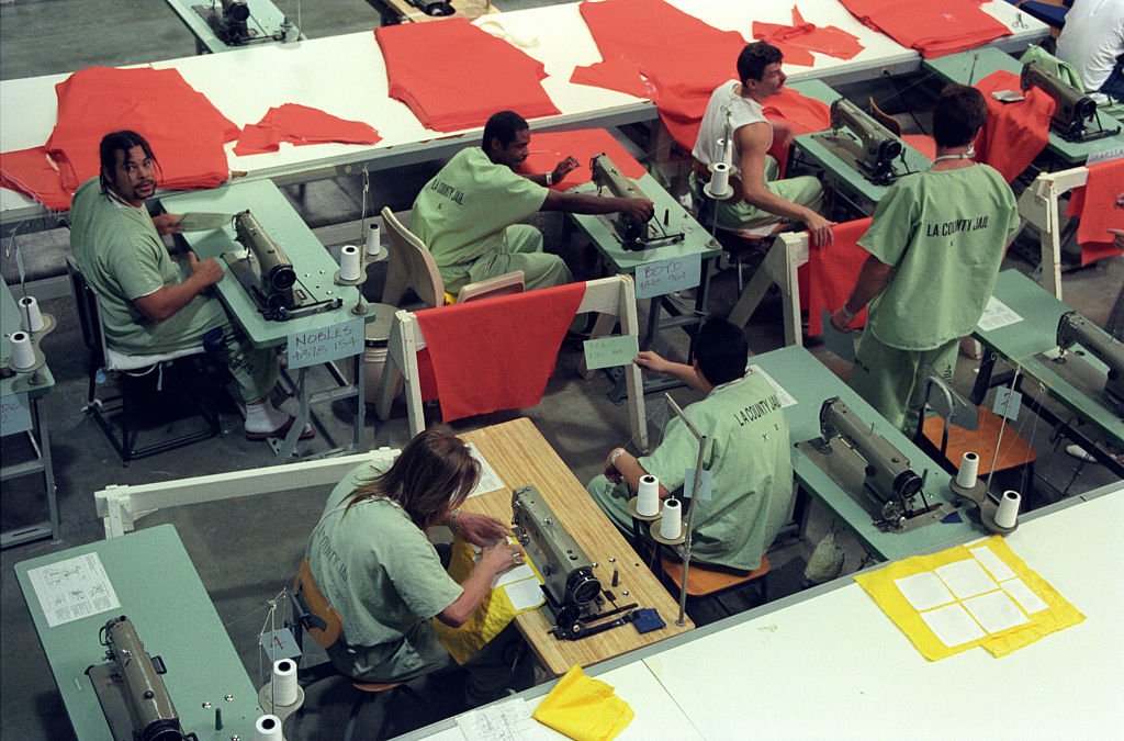 Inmates in green uniforms work at sewing stations, crafting red clothing pieces in a large, organized workshop. The image showcases a prison labor program