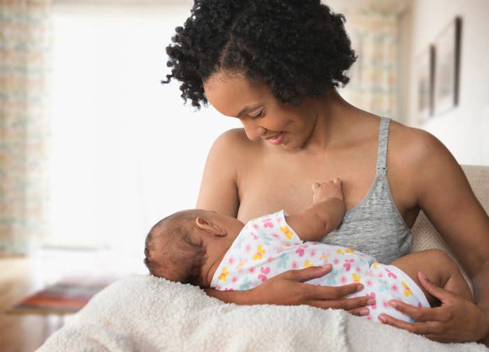 A woman is breastfeeding a baby who is wearing a onesie with a floral pattern. Both appear to be indoors in a warm, softly lit room