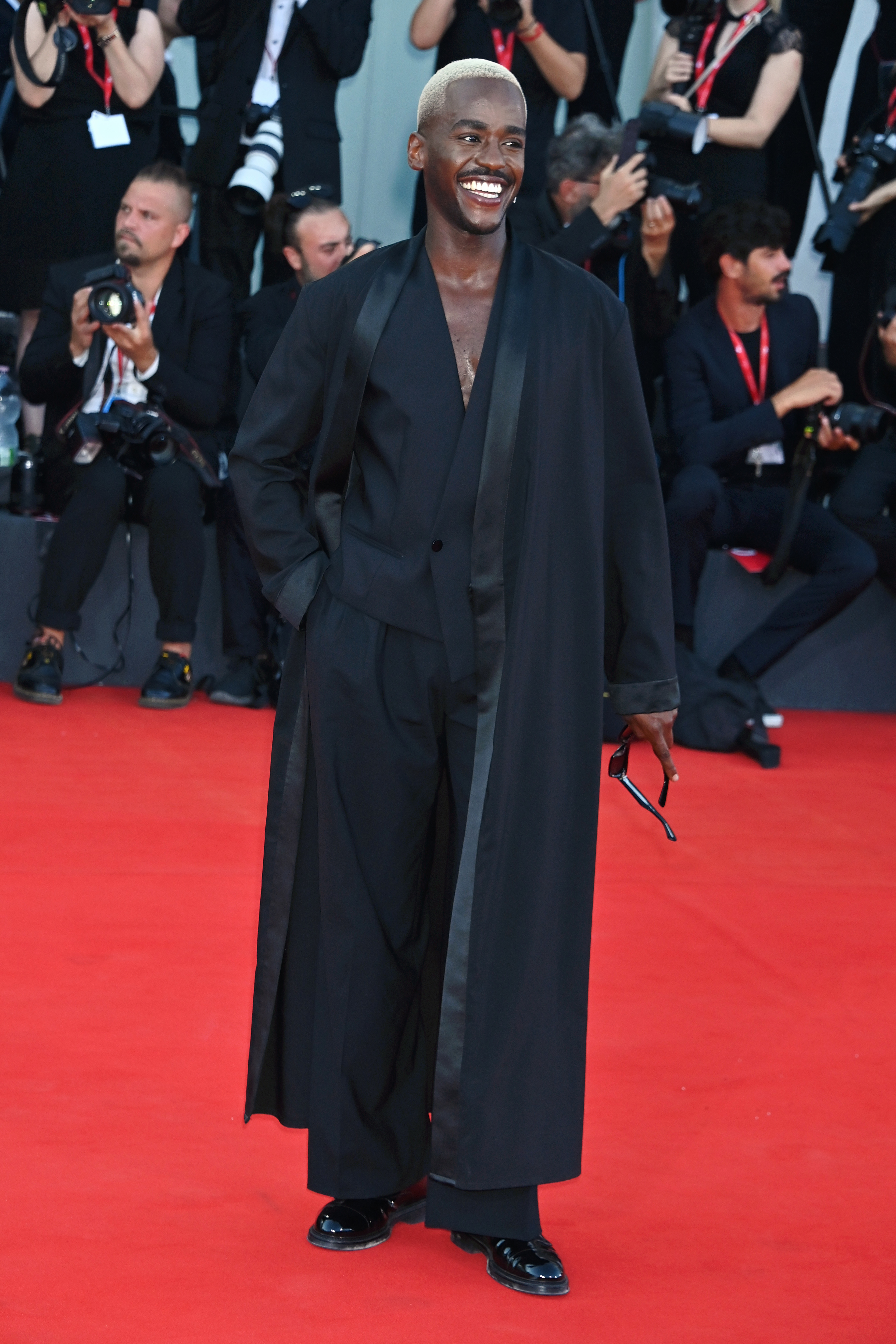 Ncuti Gatwa in a suit with a long overcoat and holding sunglasses, smiling on the red carpet with photographers in the background