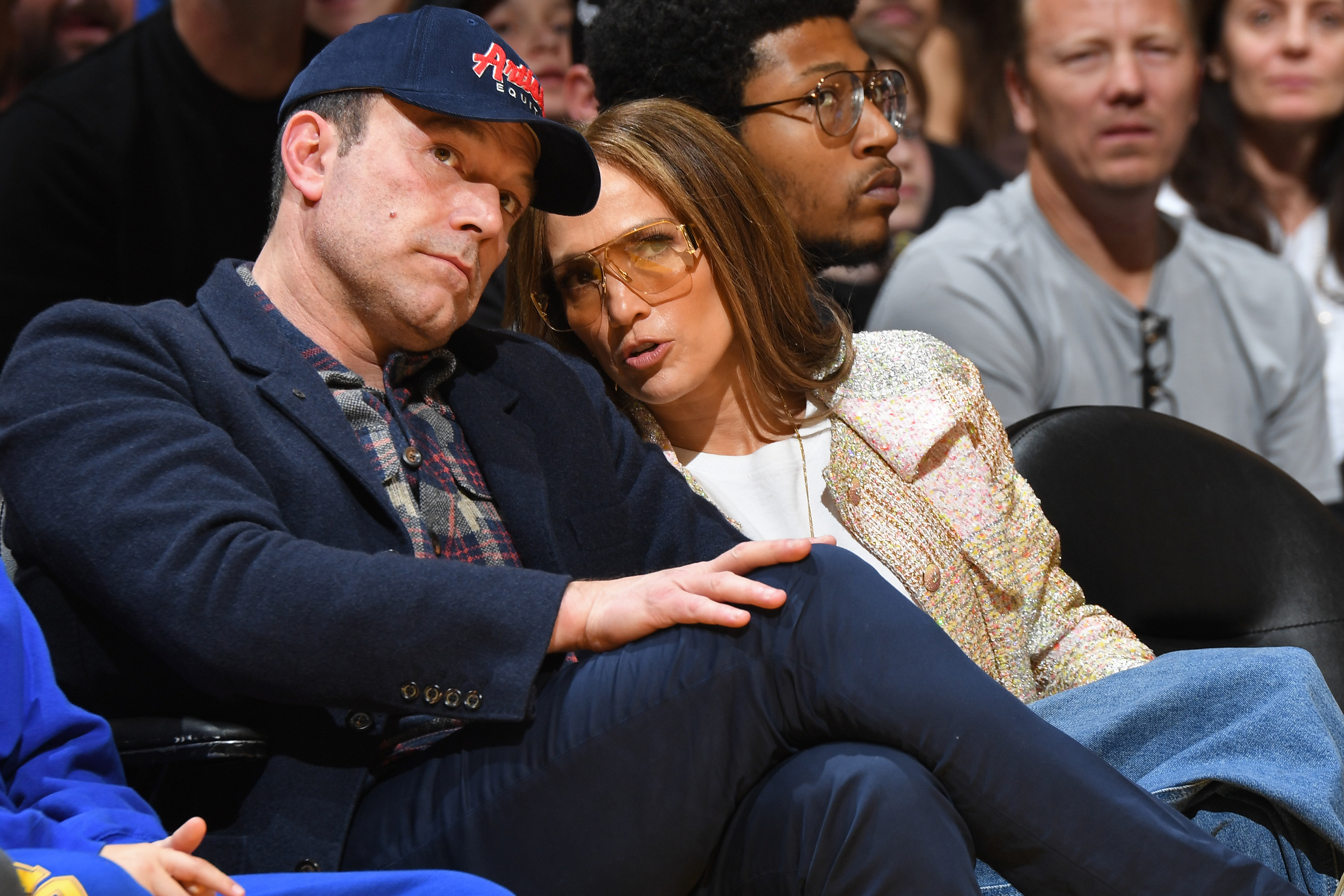 Ben Affleck and Jennifer Lopez sit closely together at a sports event. Ben wears a cap and jacket; Jennifer wears a patterned coat and large sunglasses